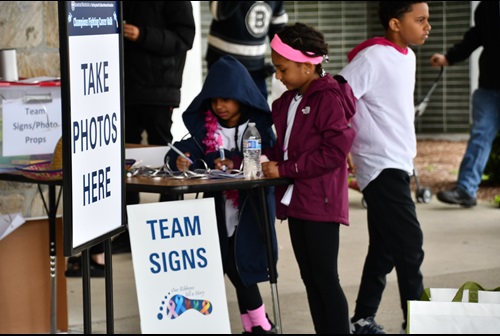 2 little girls approx. between the ages of 5-8 look pleasant, content, and concentrated as they appear to be creating a team sign at a table marked "team signs", an additional sign is visible offering participant photos, another male child passes just behind them