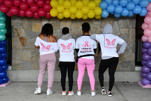 A team of 4 women stand in front of a concrete wall adorn with a balloon wreath, on the back of their matching t-shirts a pink butterfly is spread across with the names of loved ones printed too small for readability, the women have their arms drapped across eachothers shoulders 